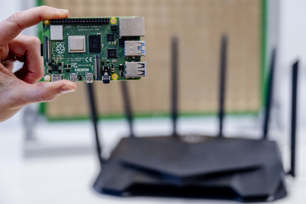Cette carte contient une surface contrôlée par logiciel, une Surface Intelligent Reconfigurable (RIS). Cette technologie permet de canaliser électromagnétiquement les signaux afin de les recevoir au mieux. Une main tenant une carte Raspberry Pi devant un routeur.